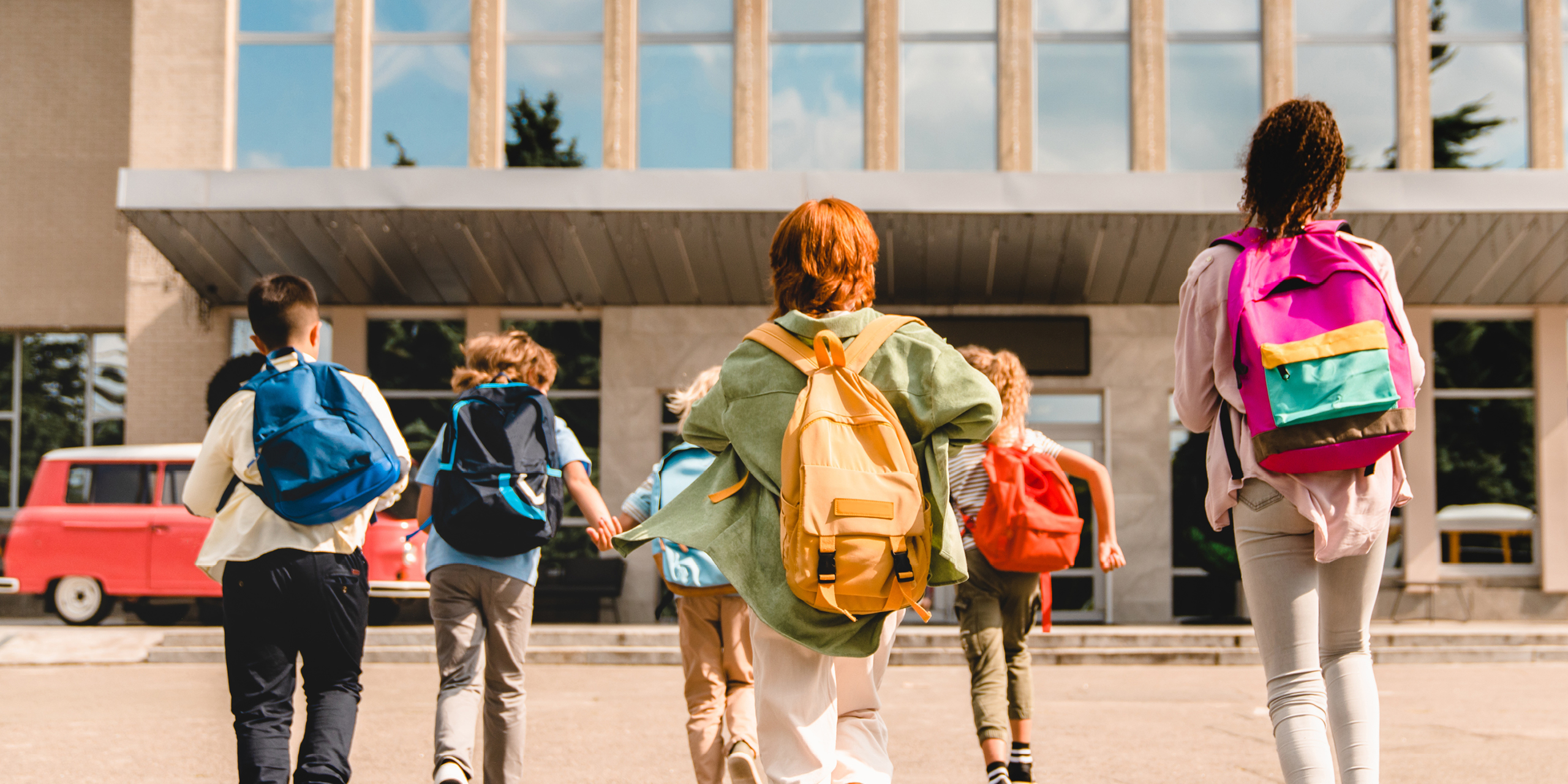 A diverse group of children walking across a street to the entrance of a school with backpacks on.