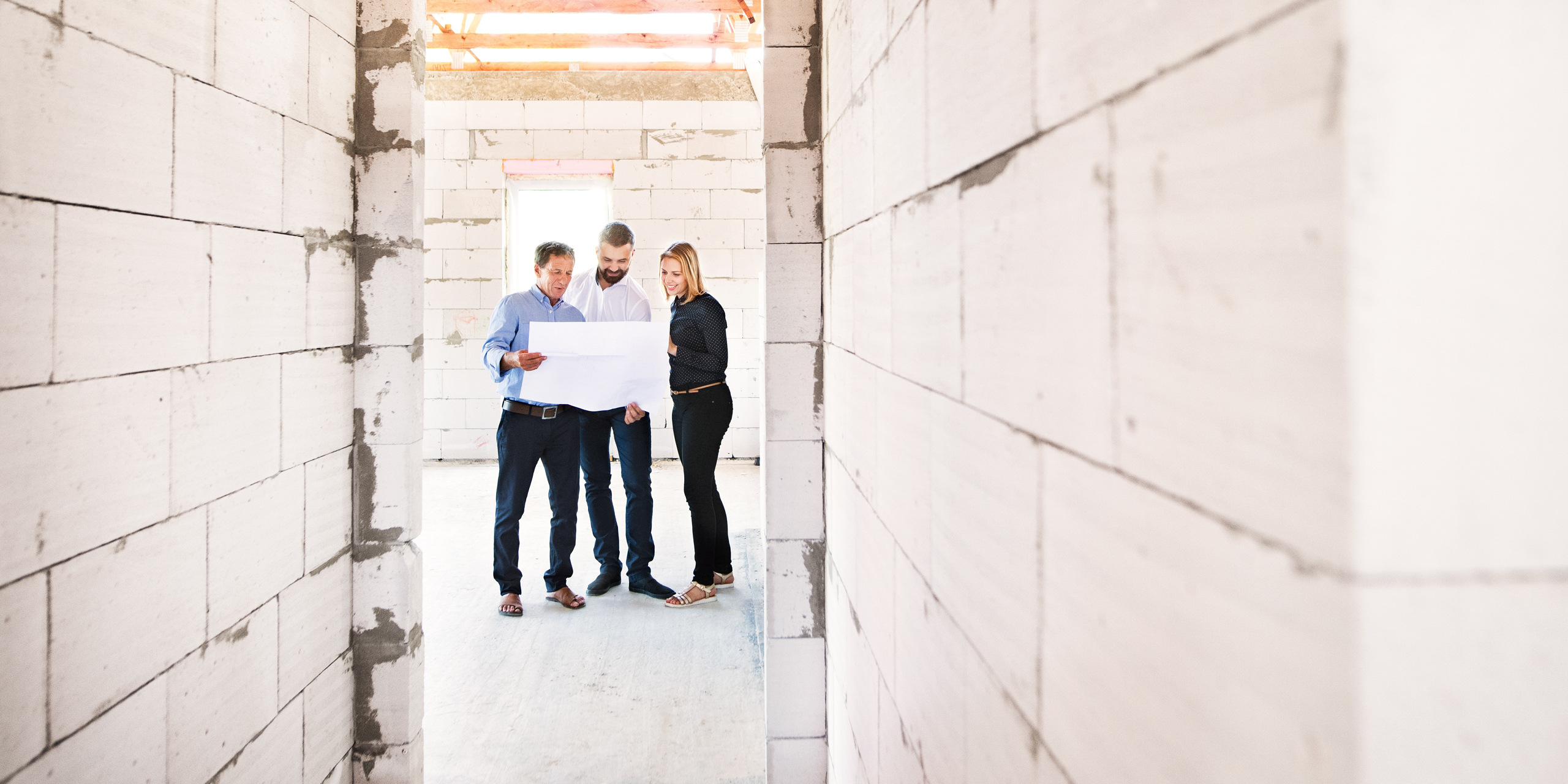 A man and woman look at blueprints with a homebuilder in a house under construction.